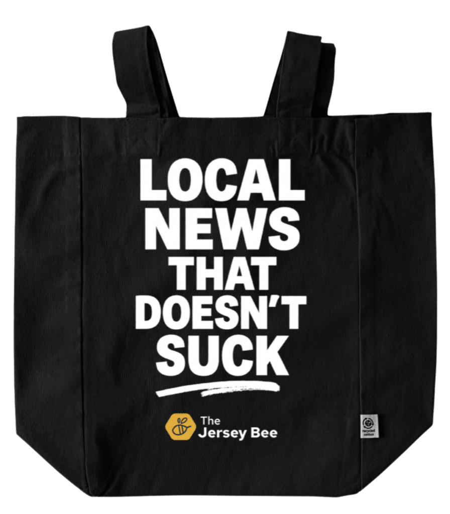 A black canvas tote bag with the words "Local news that doesn't suck" centered in white, capital letters and The Jersey Bee logo them.