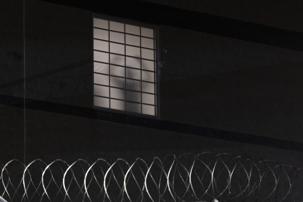 A nighttime photo of a dark facility wall. A single frosted window with a black grid of bars is pale white from interior light except for the gray silhouette of a person standing behind it. Coils of silver razor wire run across the bottom in the foreground.