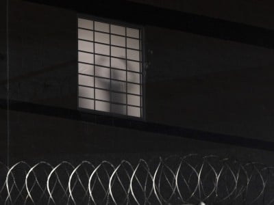 A nighttime photo of a dark facility wall. A single frosted window with a black grid of bars is pale white from interior light except for the gray silhouette of a person standing behind it. Coils of silver razor wire run across the bottom in the foreground.
