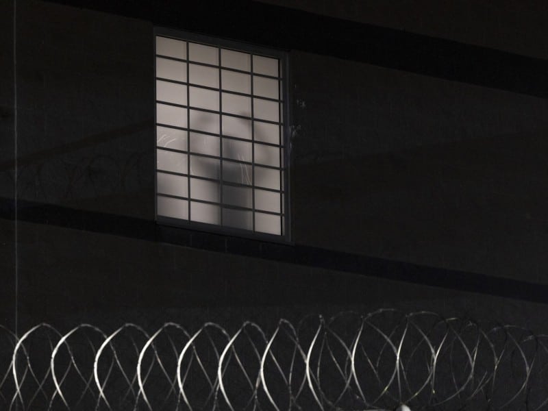 A nighttime photo of a dark facility wall. A single frosted window with a black grid of bars is pale white from interior light except for the gray silhouette of a person standing behind it. Coils of silver razor wire run across the bottom in the foreground.